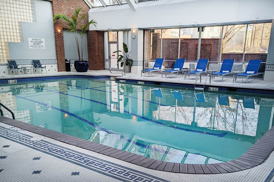 Indoor heated pool with blue loungers, brick walls, skylights, and glass enclosure