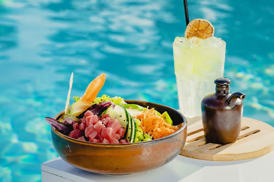 Poke bowl and cocktail served poolside with turquoise water background