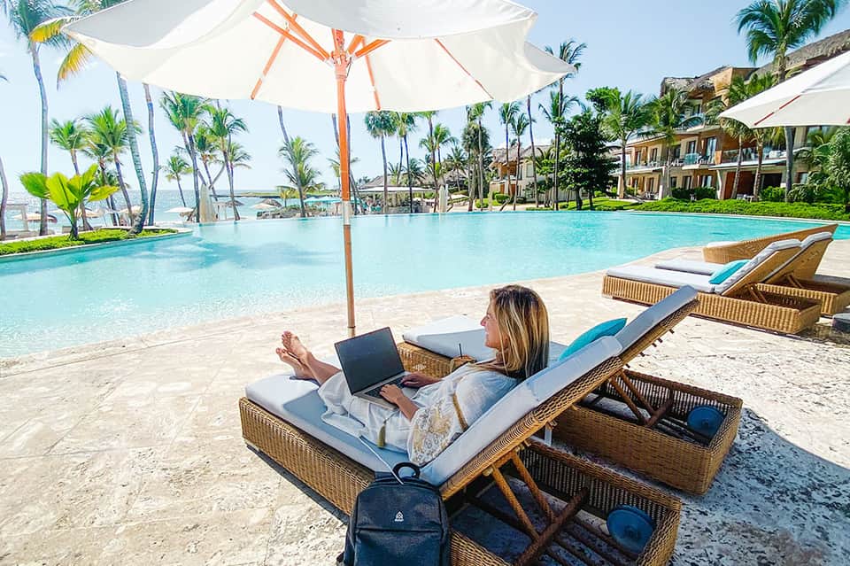 Woman working on laptop on lounge chair under large umbrella by infinity pool with palm trees