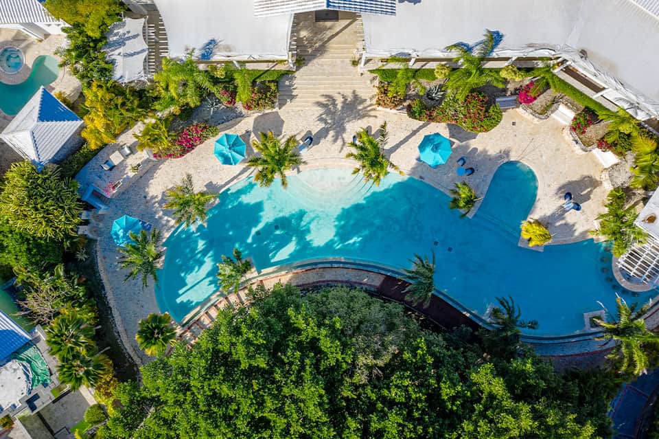 Aerial view of tropical resort pool with palm trees, blue umbrellas, and sandy beach area surrounded by lush landscaping