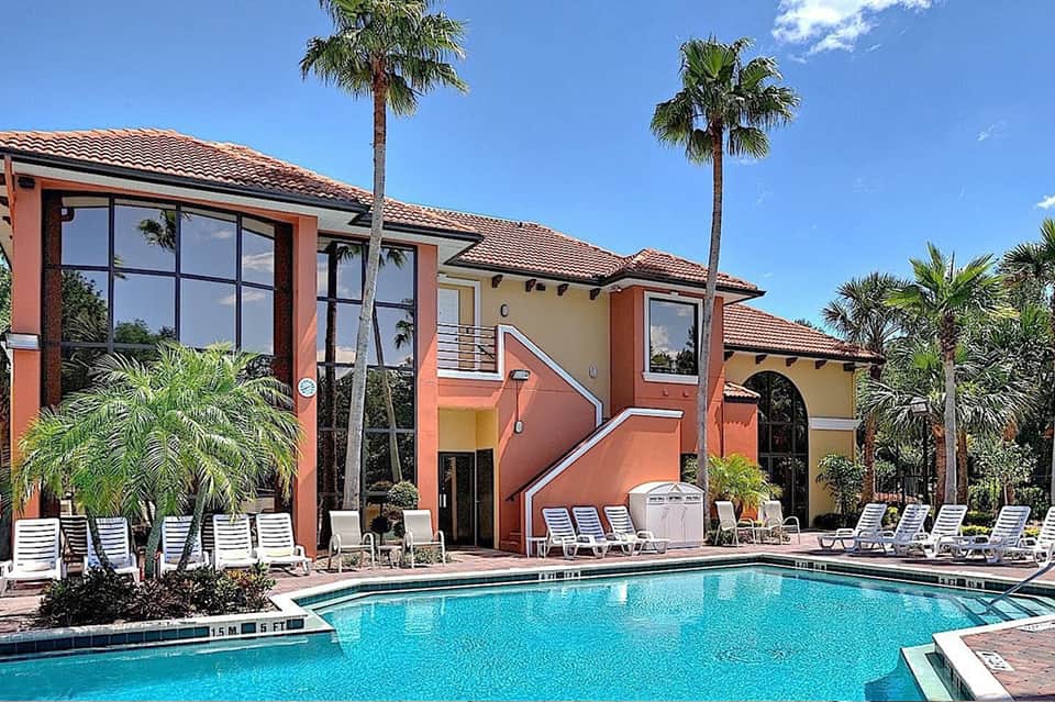 Resort pool area with coral-colored buildings, palm trees, white loungers, and clear blue sky