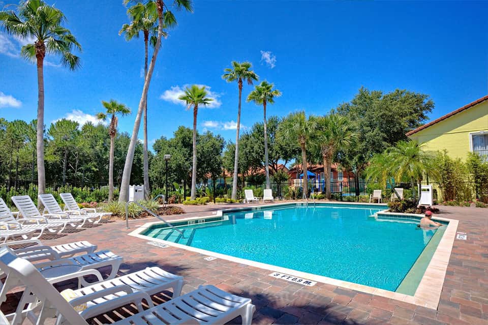 Resort pool surrounded by tall palm trees, white lounge chairs, brick patio, and tropical landscaping