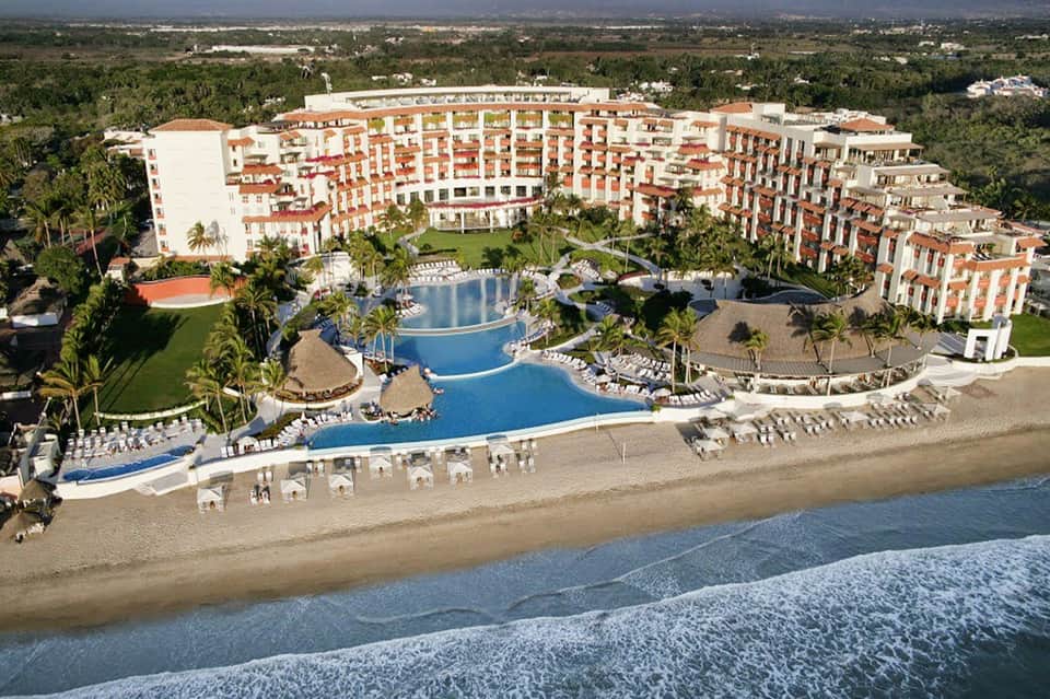 Aerial view of large beachfront resort with curved buildings, multiple pools, and sandy beach
