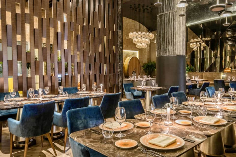 Fine dining restaurant with blue velvet chairs, geometric wood wall panels, crystal chandeliers, and elegant place settings