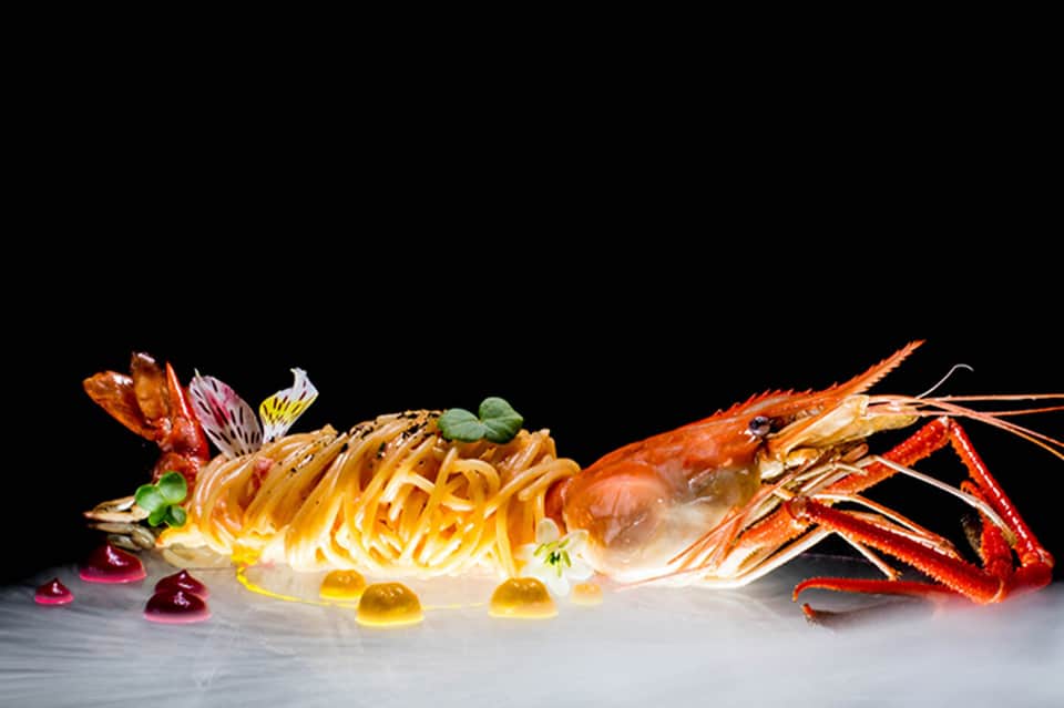 Elegant plated lobster pasta dish with decorative chocolate elements and colorful garnishes on white plate