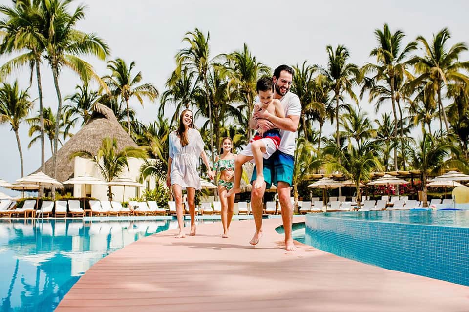 Family of four jogging on a dock between two pools at a tropical resort with palm trees and thatched roofs