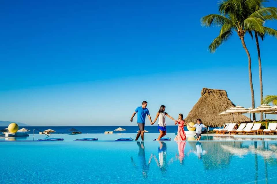 Family playing in shallow resort pool with ocean, palm trees, and tiki hut background