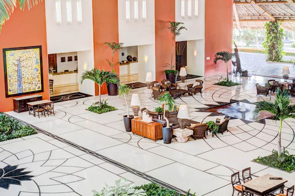 Spacious hotel lobby with orange accent walls, white columns, potted plants, lounge seating, and patterned floor