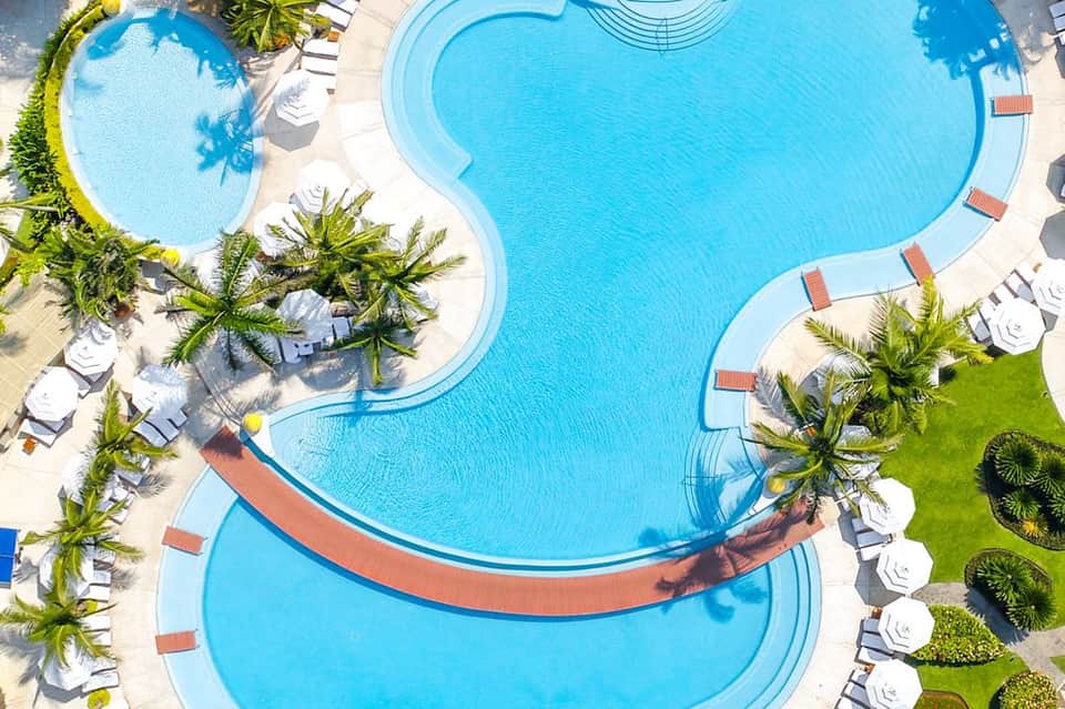 Aerial view of multi-shaped resort pool complex with palm trees, loungers, and sandy areas