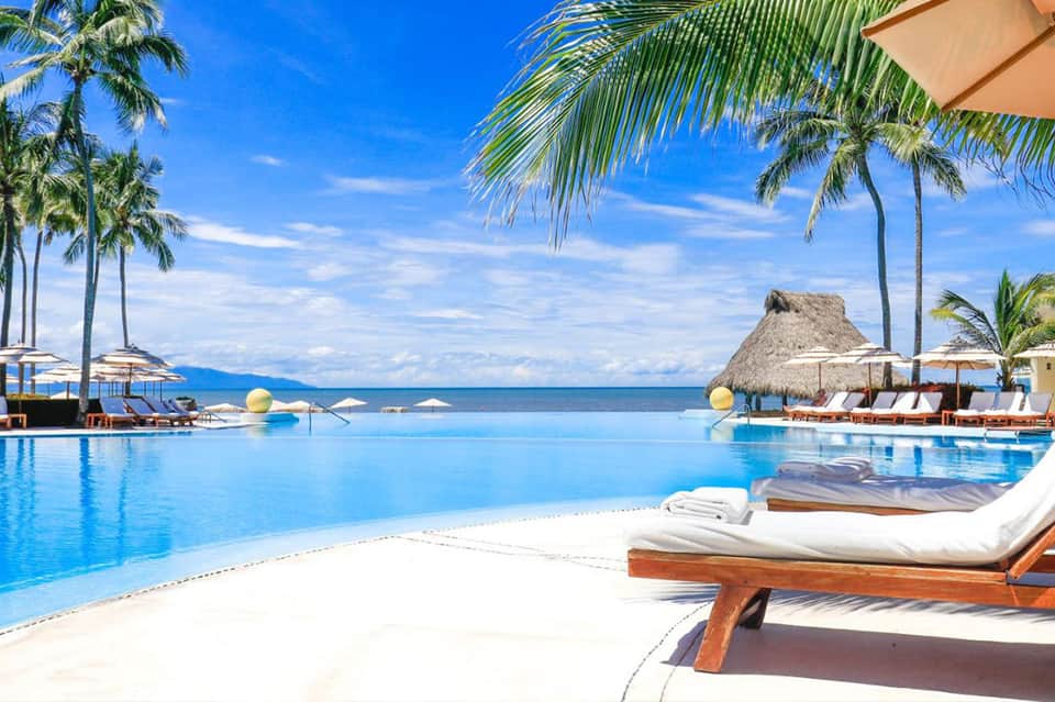 Luxury infinity pool blending with ocean horizon, white lounge beds, thatched umbrella, and palm trees at sunset