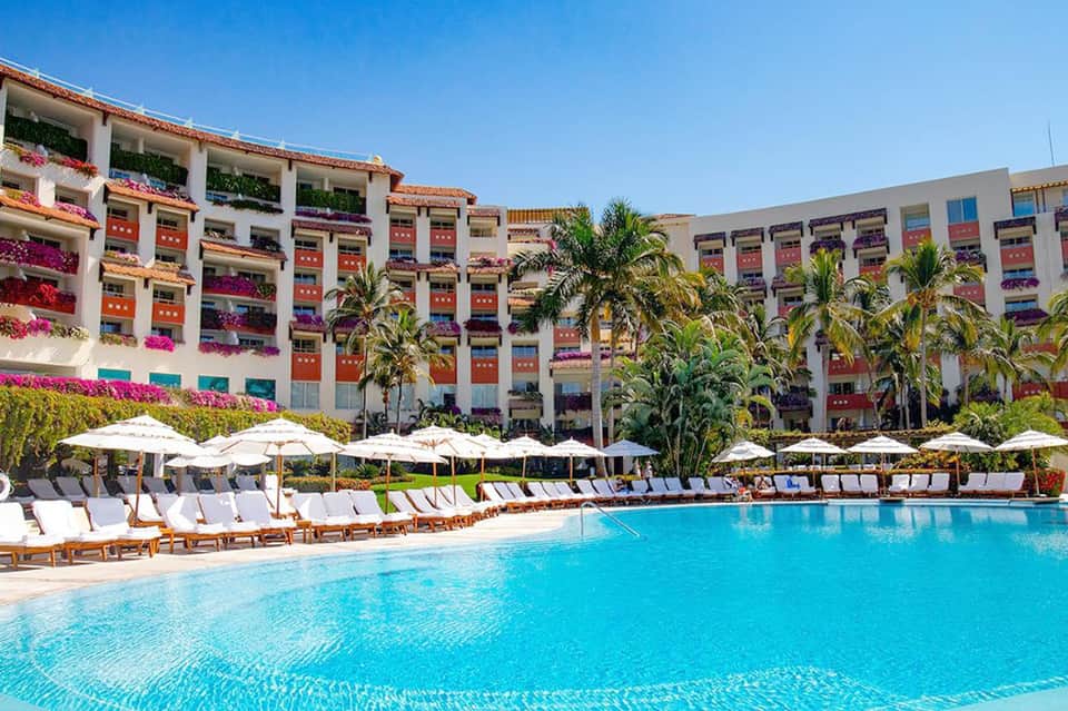 Large resort pool with turquoise water, white lounge chairs, colorful flower-draped building facade