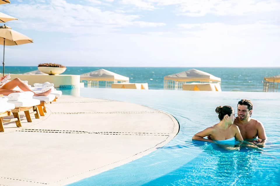 Couple swimming in infinity pool overlooking ocean with beach cabanas and umbrellas