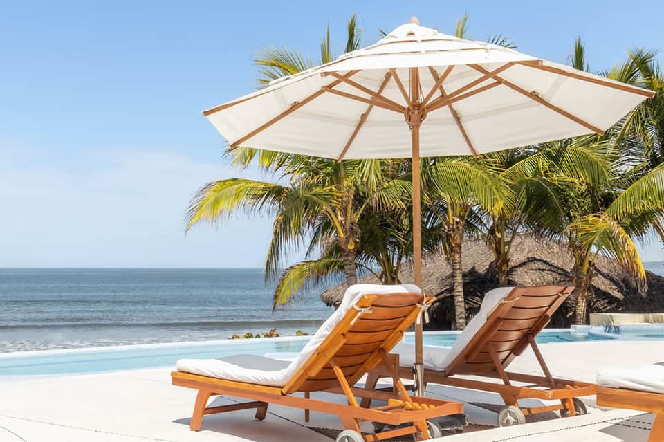 Beachfront lounge chairs with wooden frames and cream cushions under large umbrella, palm trees, and ocean view