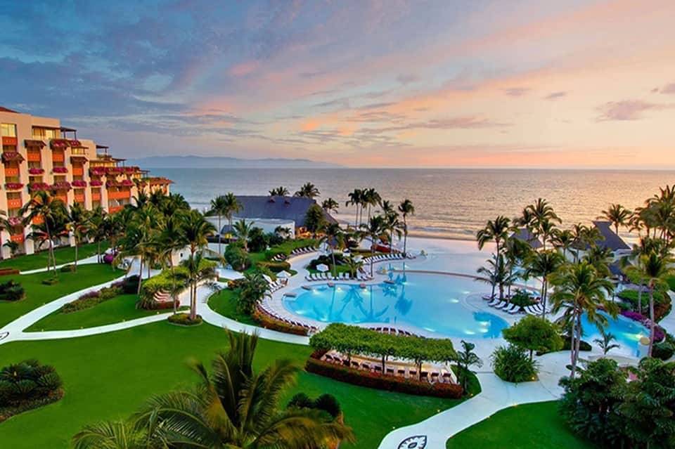 Beachfront resort aerial view with pool, palm trees, ocean sunset, and colorful buildings