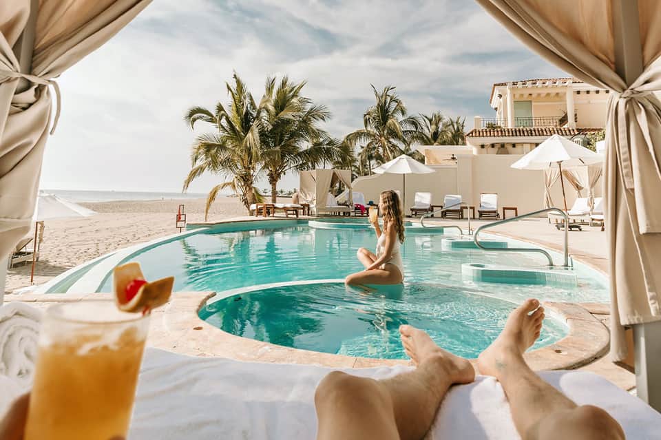 Beachfront pool with turquoise water, palm trees, white umbrellas, and ocean view beyond