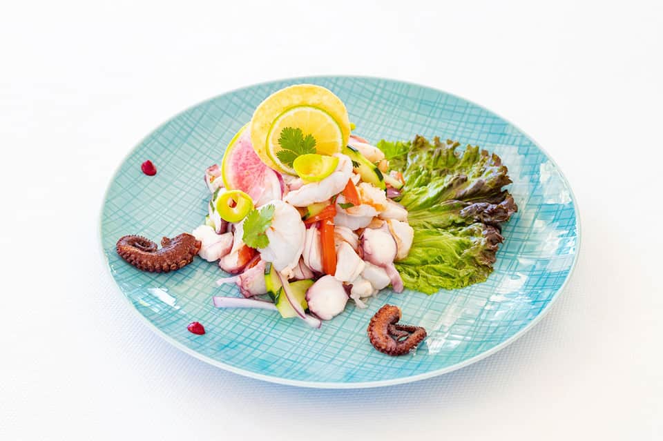 Plated octopus ceviche on blue dish with lemon, lettuce, and garnish on white background
