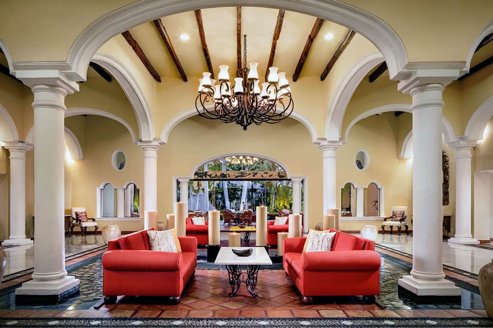 Grand hotel lobby with arched columns, crystal chandelier, red armchairs, and tropical garden views through glass doors