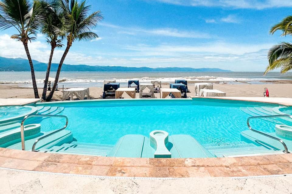 Resort pool with beach cabanas and palm trees with distant mountains and ocean backdrop
