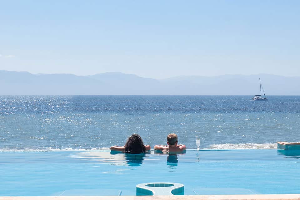 Infinity pool overlooking ocean with two swimmers, sailboat visible on water