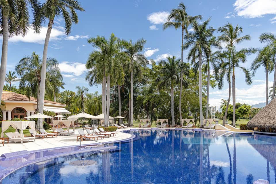 Resort pool surrounded by white cabanas, palm trees, and manicured lawn under blue sky