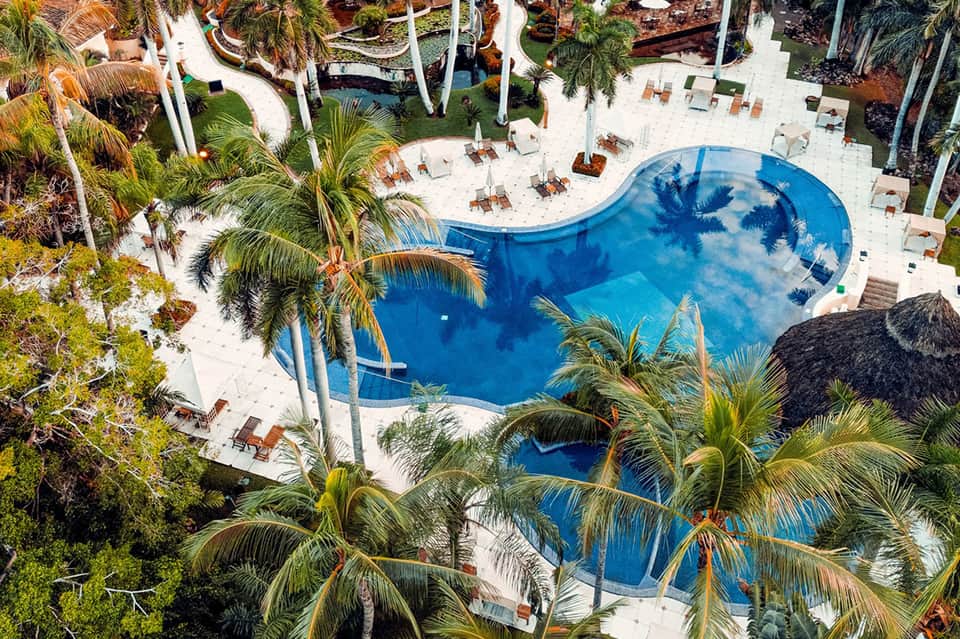 Overhead view of curved resort pool surrounded by white sand, palm trees, and loungers