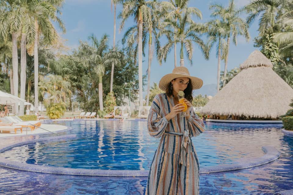 Woman in striped robe holding drink by resort pool with thatched palapa and palm trees