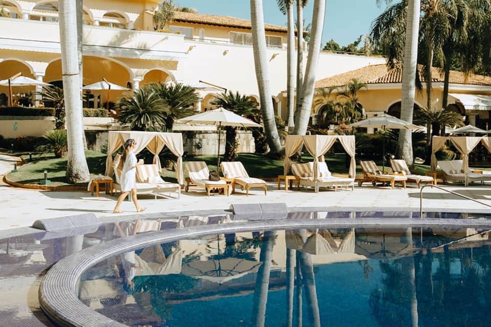 Resort pool with curved edge, striped loungers, palm trees, and yellow umbrellas