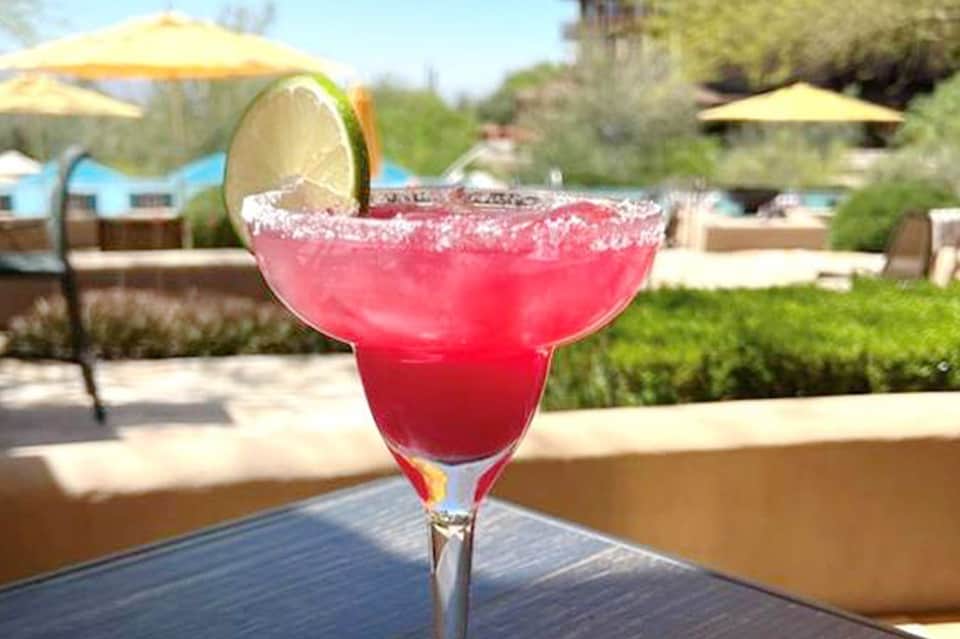 Pink margarita with sugar rim and lime garnish on poolside table with umbrellas in background