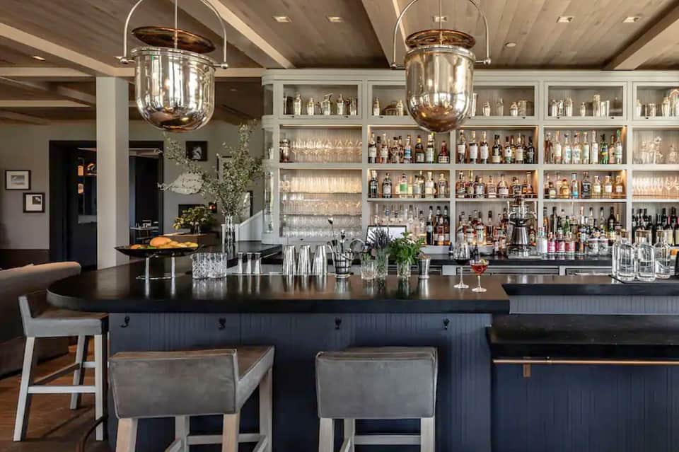 Modern bar with dark counter, pendant lighting, fully stocked shelves, and upholstered bar stools