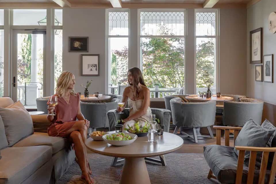 Two women enjoying drinks in an elegant lounge with floor-to-ceiling windows overlooking trees