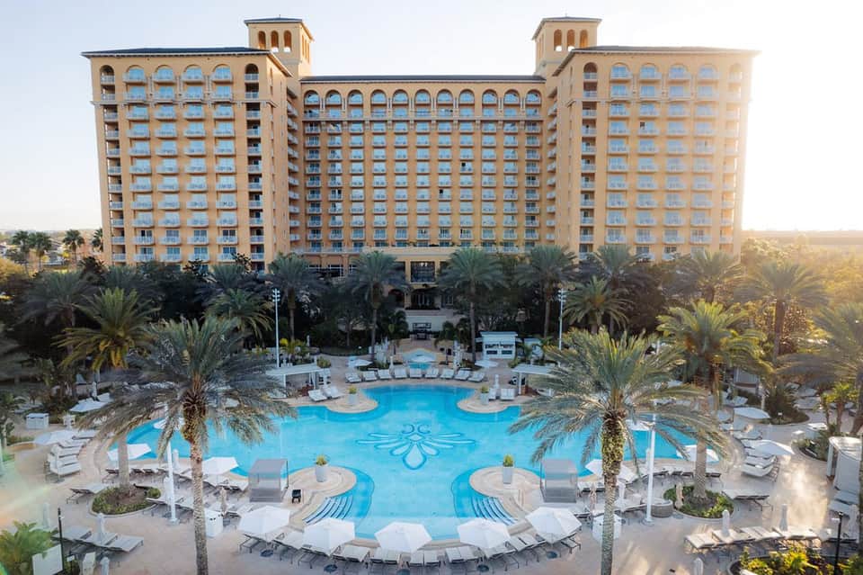 Large resort pool with ornate blue pattern, palm trees, white lounge chairs, and multi-story hotel building