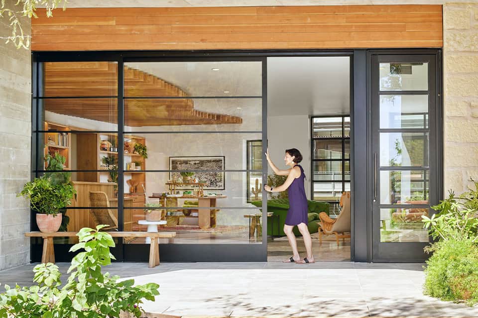 Woman opening large black-framed glass doors to a modern wooden interior with plants and shelving