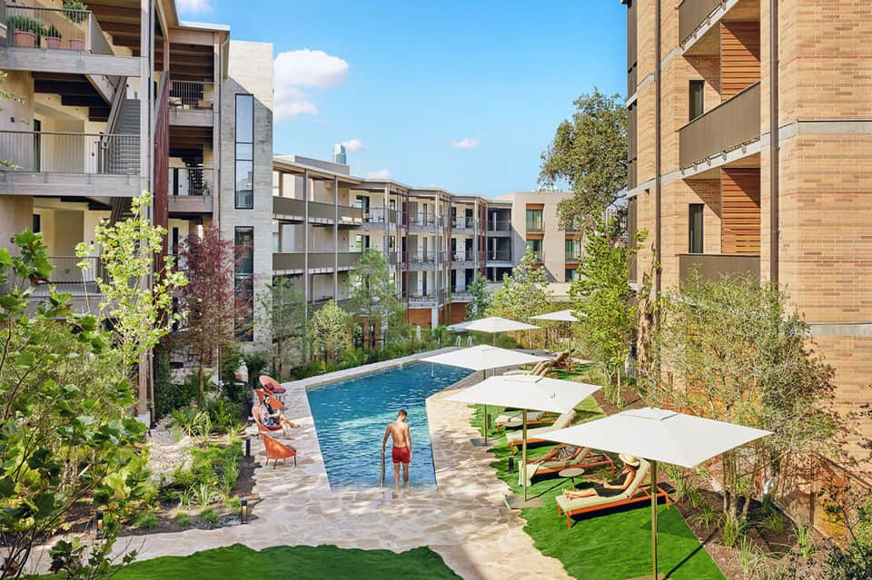 Urban resort courtyard with rectangular pool, green landscaping, white umbrellas, and surrounding modern buildings