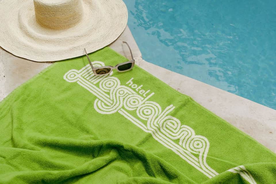 Green towel with resort logo beside sunglasses and straw hat on pool deck
