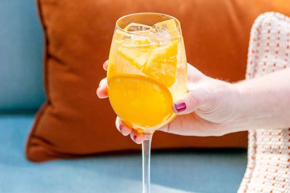 Woman's hand holding a refreshing yellow citrus cocktail with ice against colorful cushions