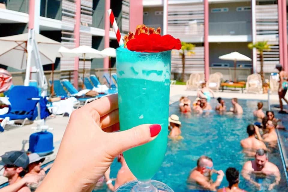 Hand holding tropical turquoise cocktail with red garnish overlooking busy resort pool area