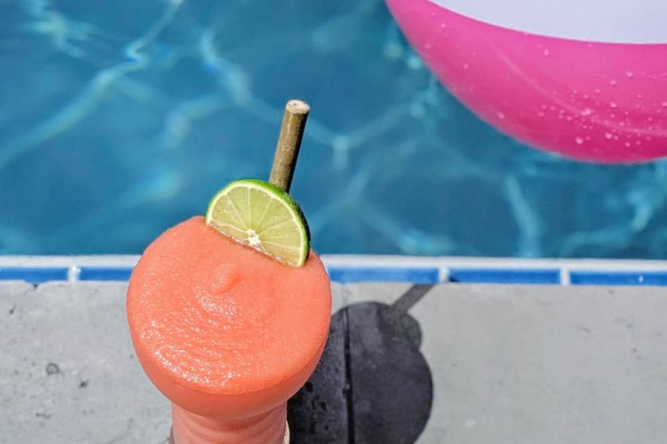 Tropical cocktail with coral-pink liquid and lime slice, poolside with floats and clear water