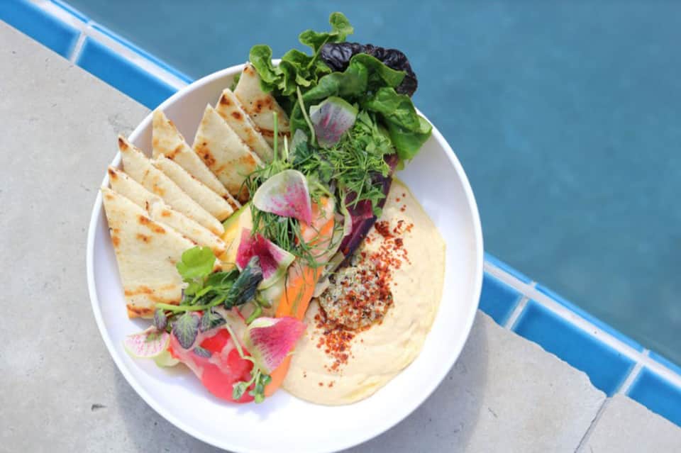 Elegant plated dish with hummus, fresh greens, colorful vegetables, pita bread, and spiced garnish by poolside