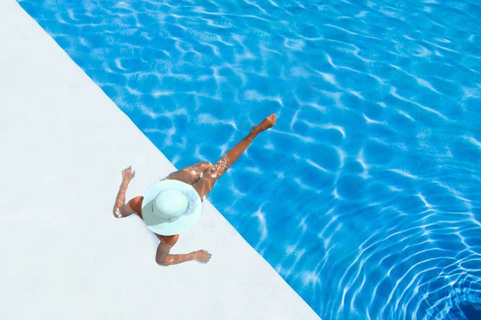 Person swimming in crystal-clear blue pool wearing white hat, overhead view