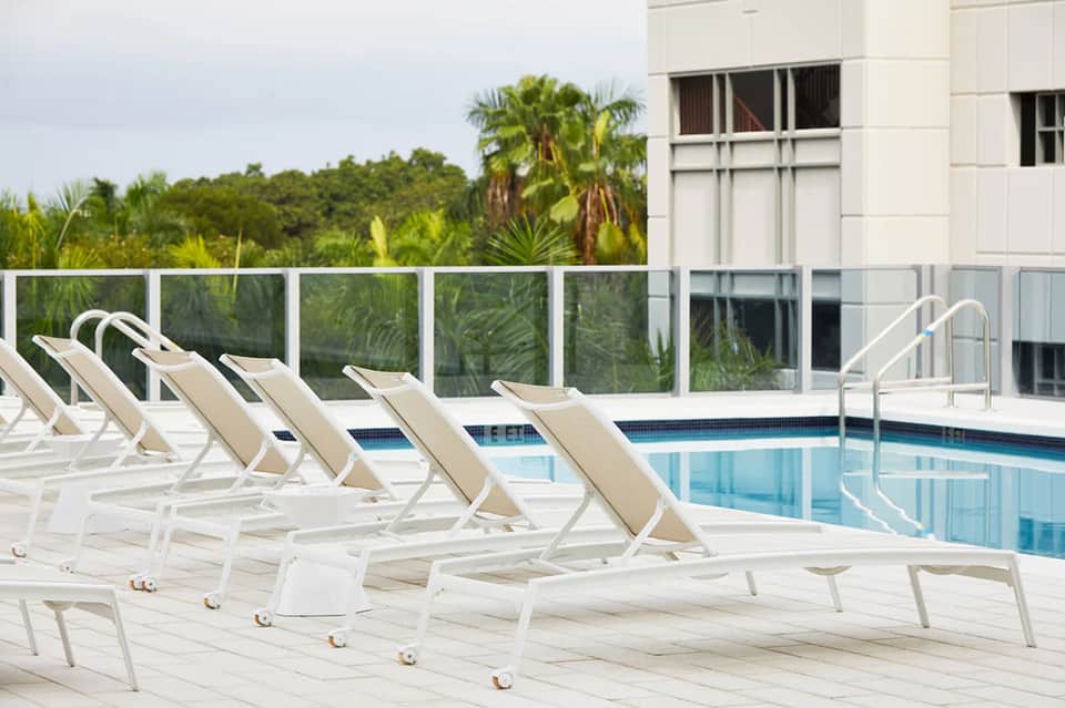 Resort pool deck with white lounge chairs, handrail, forested hillside, and modern building