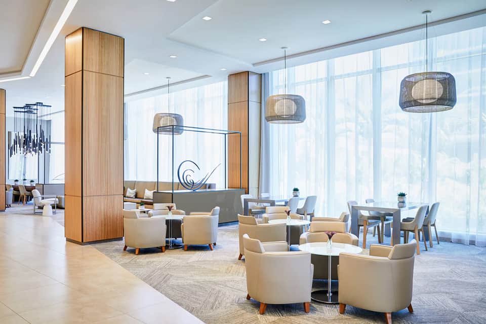 Modern hotel lobby lounge with high ceilings, wooden columns, pendant lights, beige chairs, and floor-to-ceiling windows