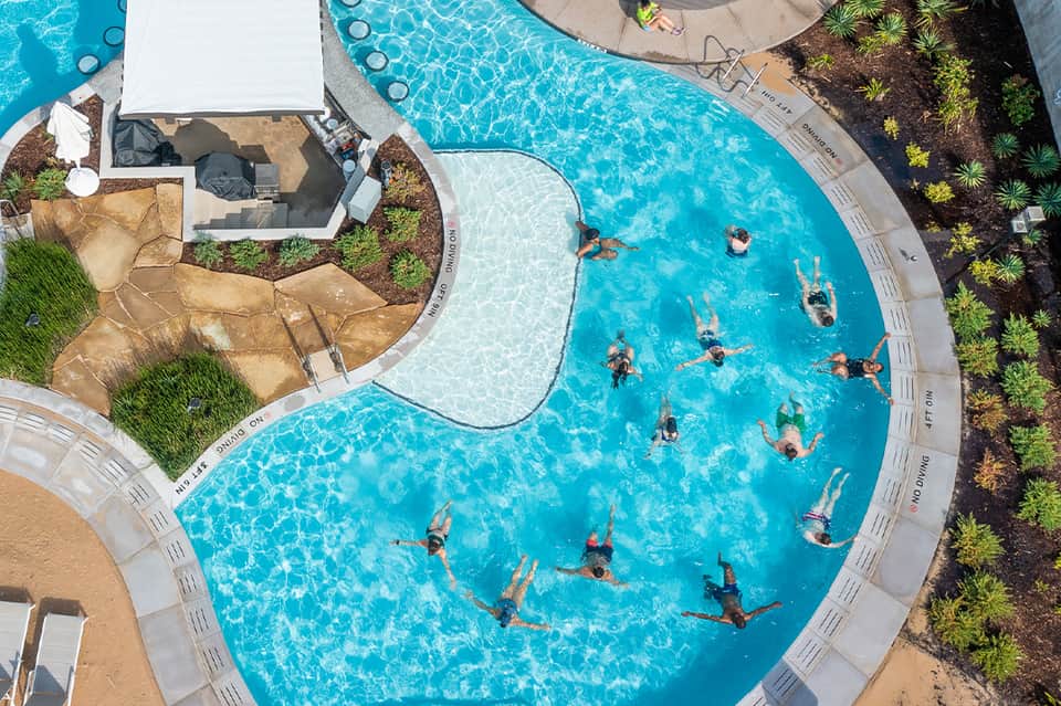 Overhead view of curved resort pool with swimmers, white umbrellas, and landscaped grounds