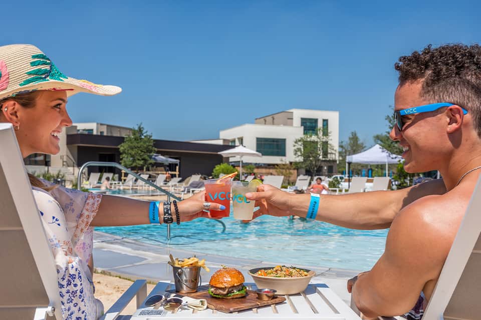 Two people toasting drinks poolside with food on the deck, modern resort architecture visible in background