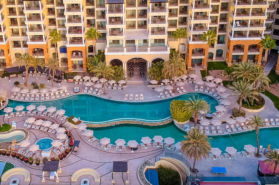 Aerial view of resort with curved pool, lounge chairs, palm trees, and orange building wings
