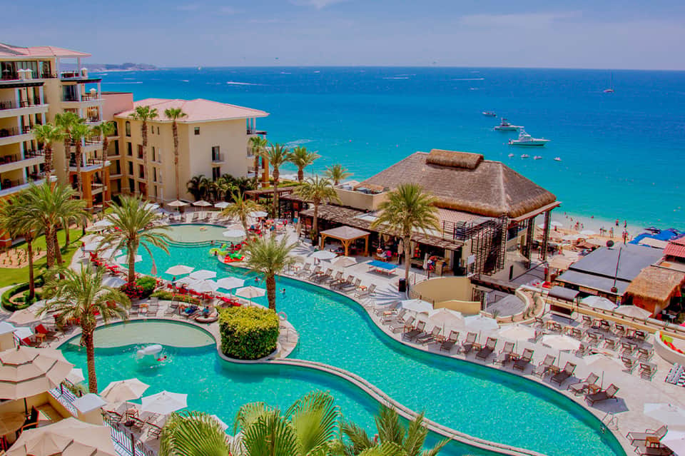 Aerial view of beachfront resort with pools, thatched cabanas, and turquoise ocean