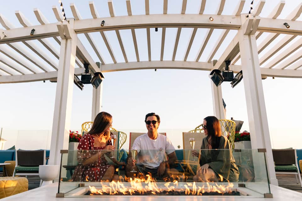 Three people sitting by outdoor fire pit under pergola at dusk