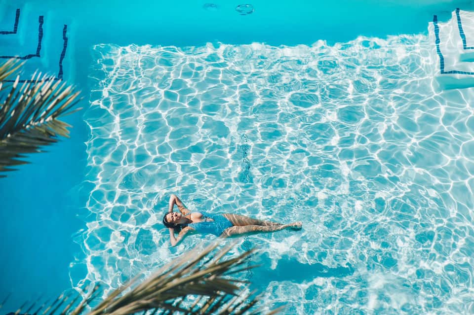 Aerial view of woman floating in crystal-clear turquoise pool water with palm fronds visible