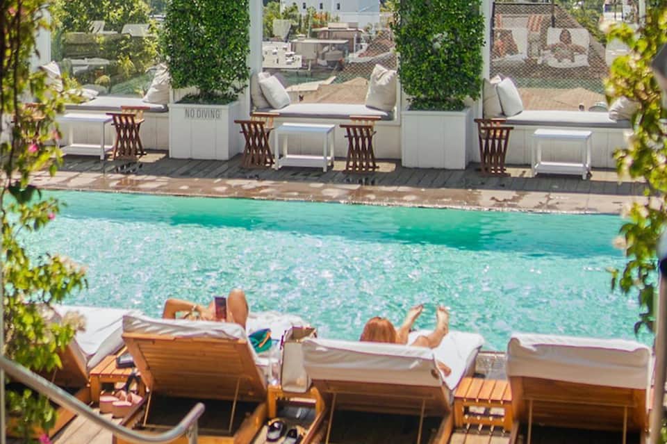 Resort pool with turquoise water, wooden lounge chairs with white cushions, and waterfront cabanas