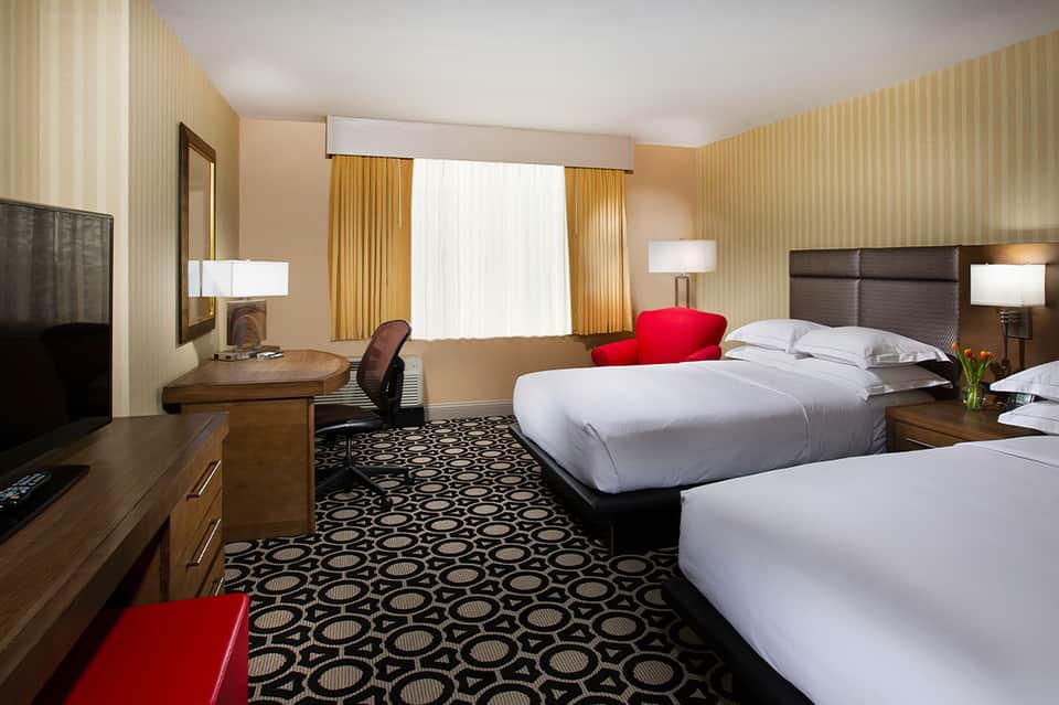 Hotel room with two queen beds, wood furnishings, patterned carpet, and warm ambient lighting