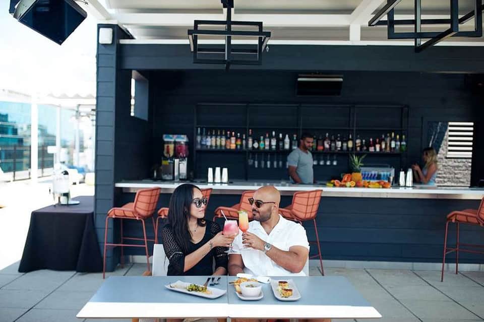Contemporary rooftop bar with couple dining, dark counter, orange seating, and city skyline backdrop